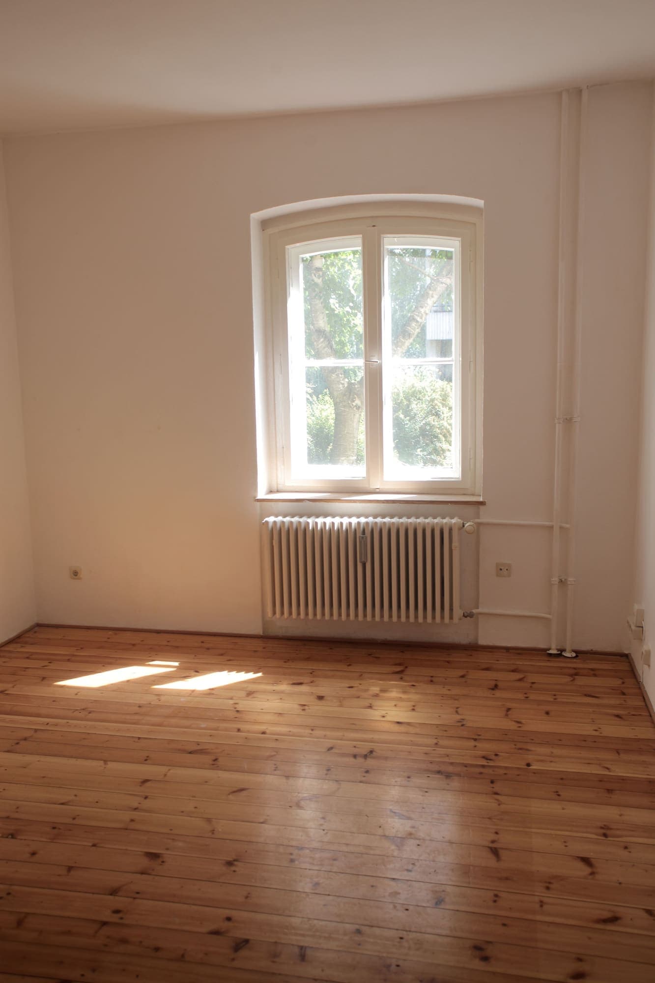 Habitación vacía de un piso con suelo de madera, radiador y una ventana con luz natural